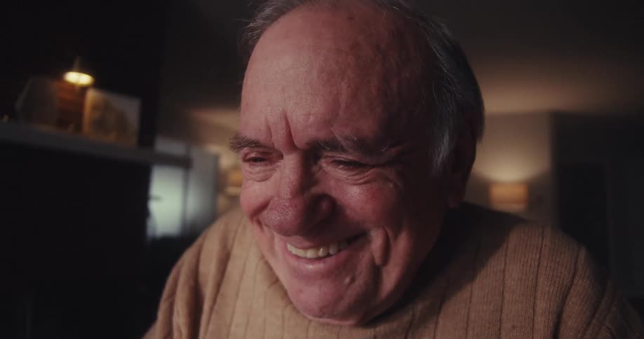 An elderly man sitting alone in a warmly lit room smiles with visible emotion while on a video call