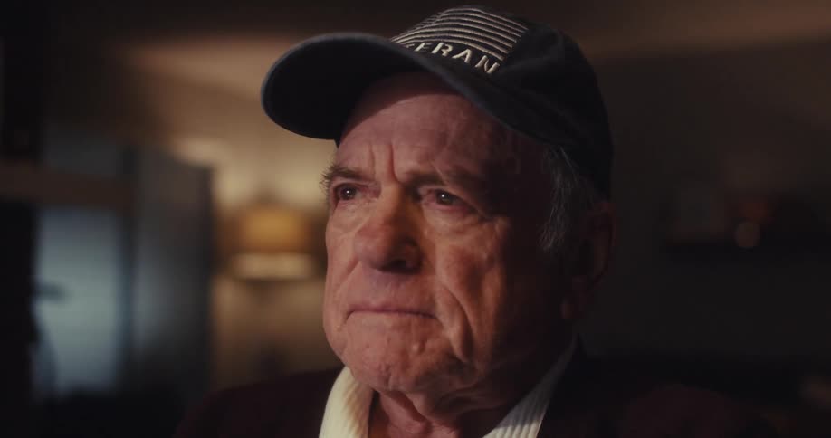 An elderly man in a veteran cap sits indoors appearing thoughtful and slightly sad