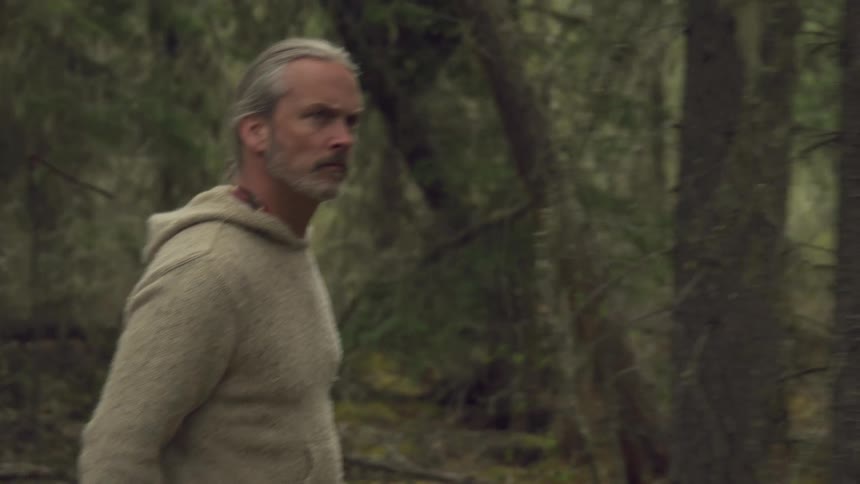 A man in a beige sweater walks through a forest with a hunting rifle alert and watchful