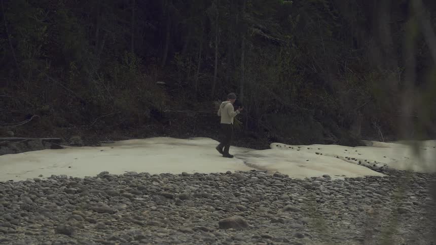 A man is walking near a riverbed holding rocks and stems in his hands surrounded by a forested area