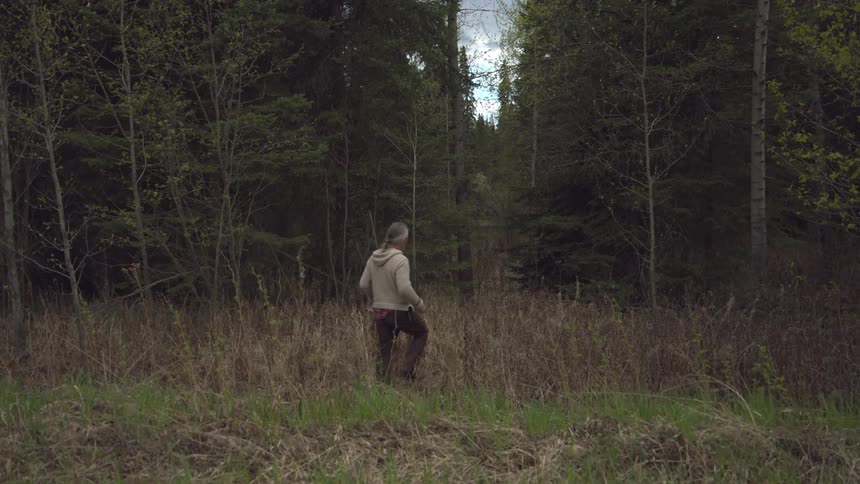A man carrying a plastic tub walks through a grassy field surrounded by dense forest