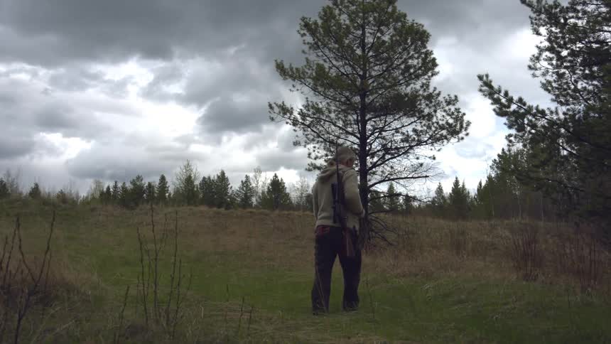 A person with a rifle slung over their shoulder walks across a grassy field under a cloudy sky