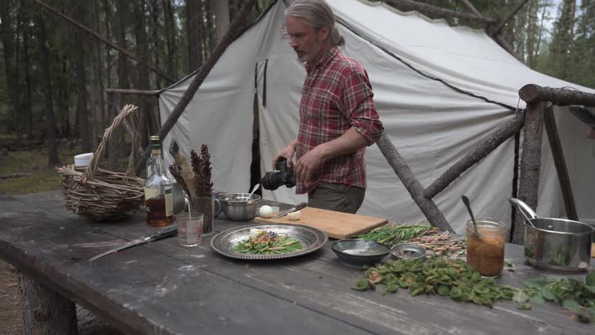 Two men photograph a rustic foraged meal at a forest campsite showcasing outdoor cooking