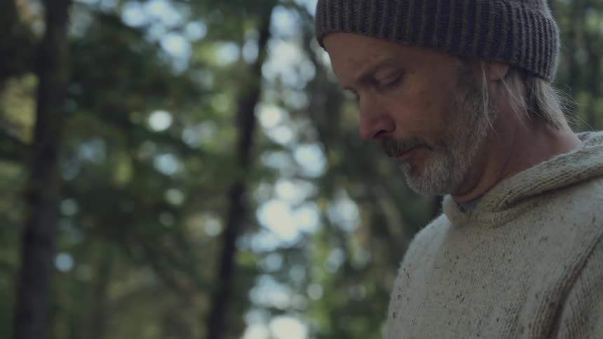 A man with a beard and beanie looks down pensively in a forest setting