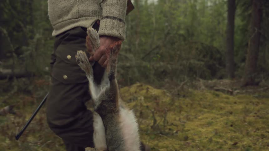 A man walks through a forest holding a rifle and a recently hunted animal by its legs