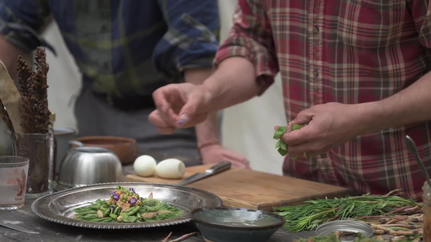 A person in a red and black plaid shirt meticulously garnishes a plate of fiddleheads with small flowers and herbs