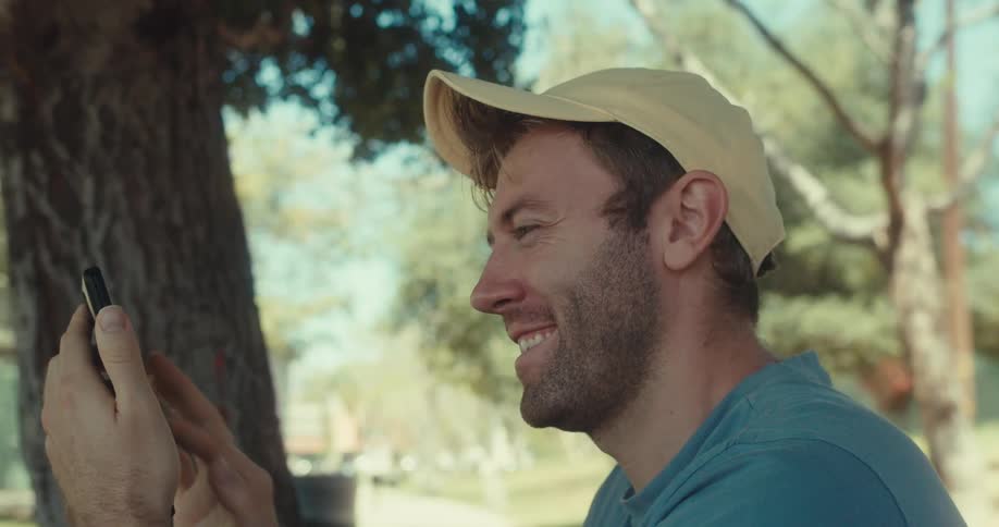 A man in a yellow cap and blue shirt sits under a tree in a park browsing his phone with a happy expression on a sunny day