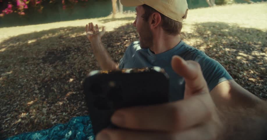 A man in a yellow cap and blue shirt sits in a park on a sunny day taking selfies with a smartphone