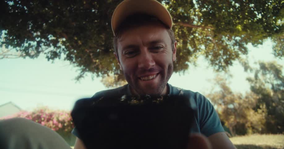 Man sitting outdoors under a tree in a field near a residential area smiles and waves during a video call on his smartphone
