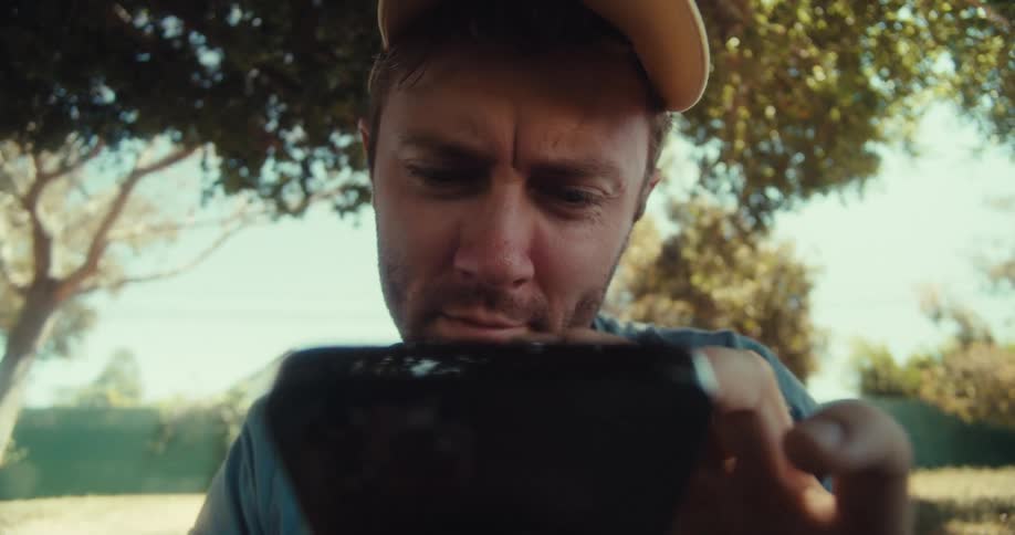 A man in a yellow cap and blue shirt sits outdoors under trees looking down while using his phone