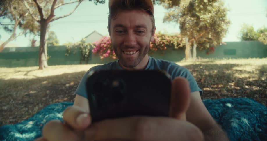 Man lies back and sits up on a blanket in a residential park while viewing his smartphone screen