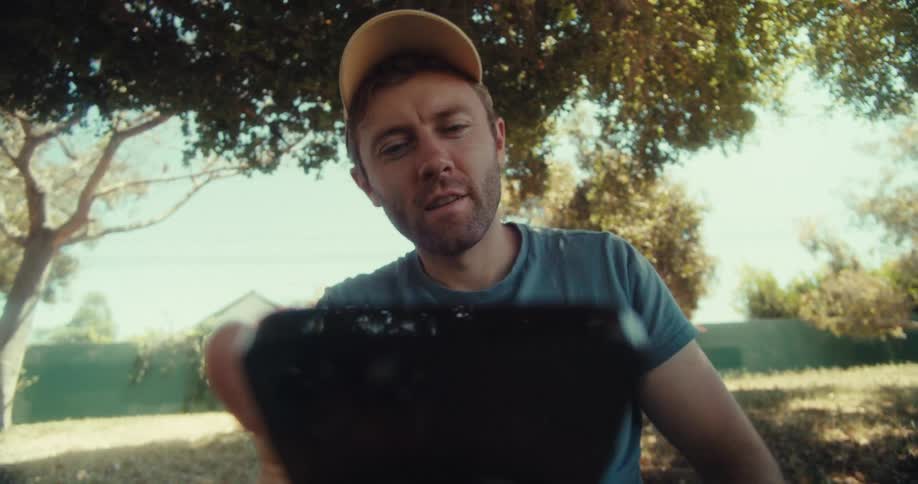 A man in a yellow cap and blue shirt sits outdoors under trees looking down while using his phone