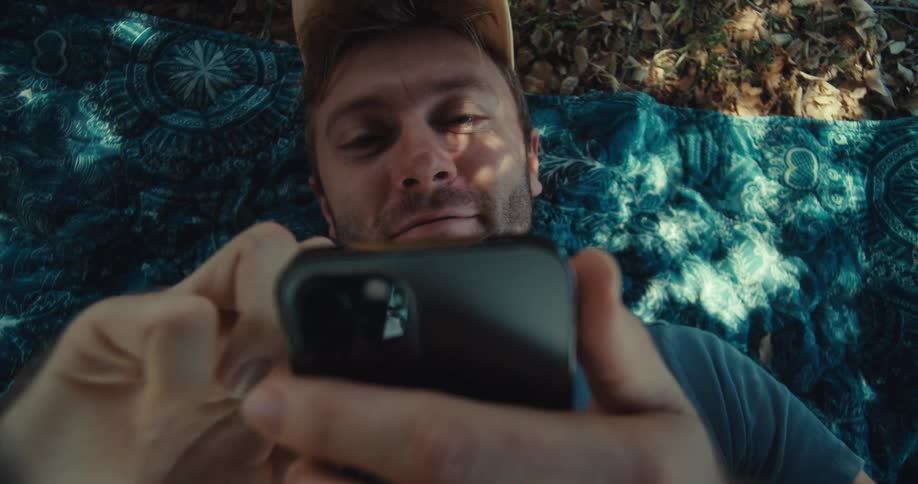 A man in a yellow cap and blue shirt sits in a park on a sunny day lies down while browsing his phone then sits back up again with a surprised and happy expression