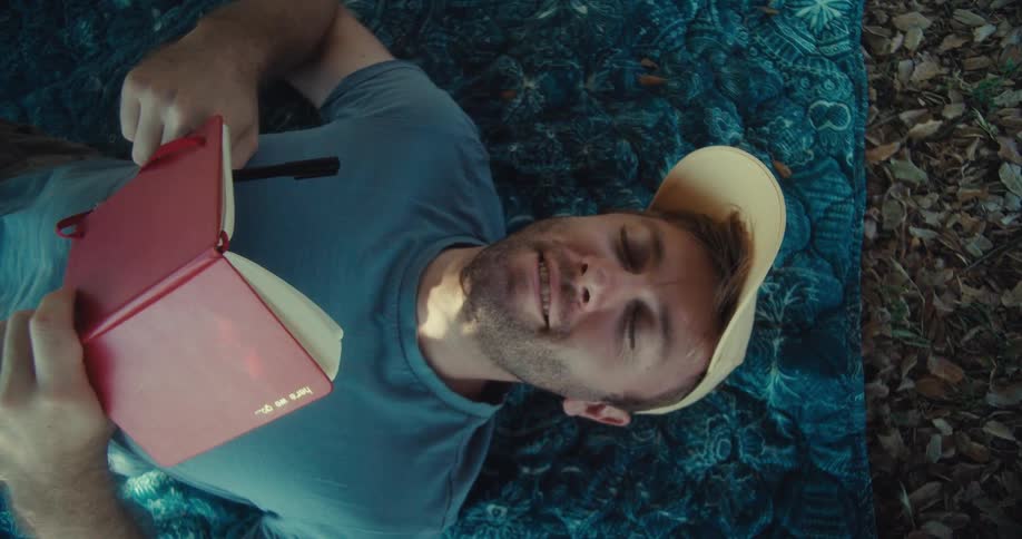 A man is laying on a blanket outside and reading a book