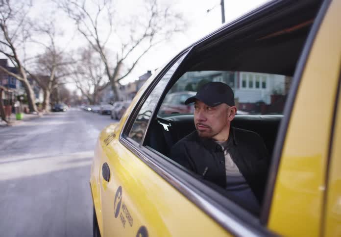 A man wearing a baseball cap looks out the window of a moving yellow taxi cab on a city street lined with parked cars and bare trees