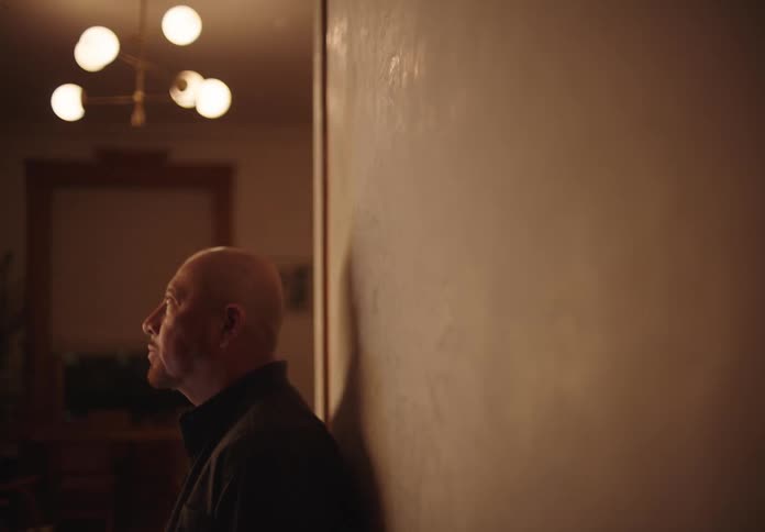 A bald man stands beside a wall inside his home deep in thought with a chandelier on the ceiling brightening the room