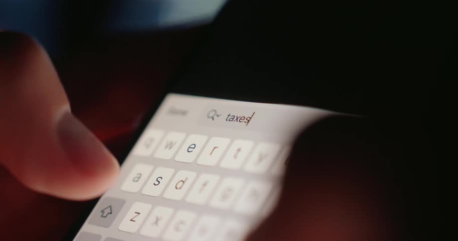 Fingers type the word taxes on a brightly illuminated smartphone keyboard in a dimly lit area