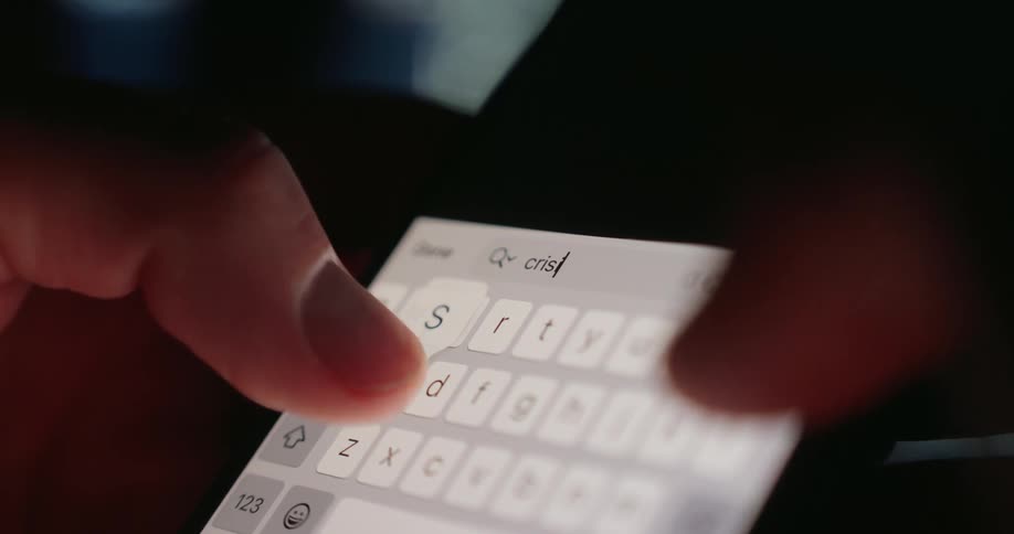 Fingers type on a brightly illuminated smartphone keyboard in a dimly lit area