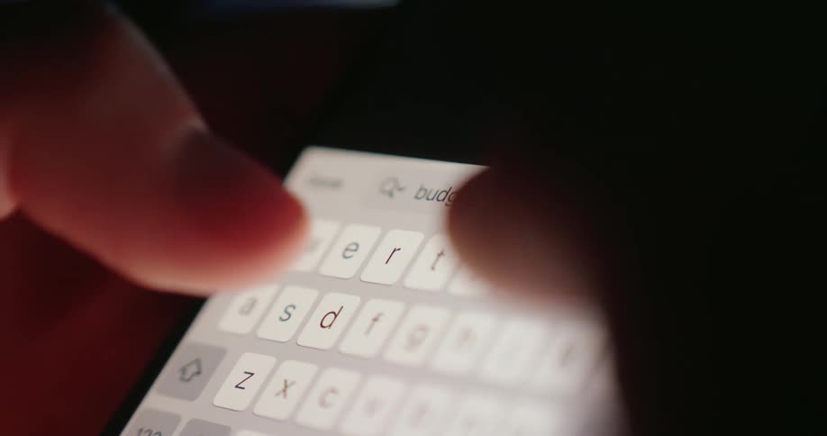 Fingers type the word budgeting on a brightly illuminated smartphone keyboard in a dimly lit area