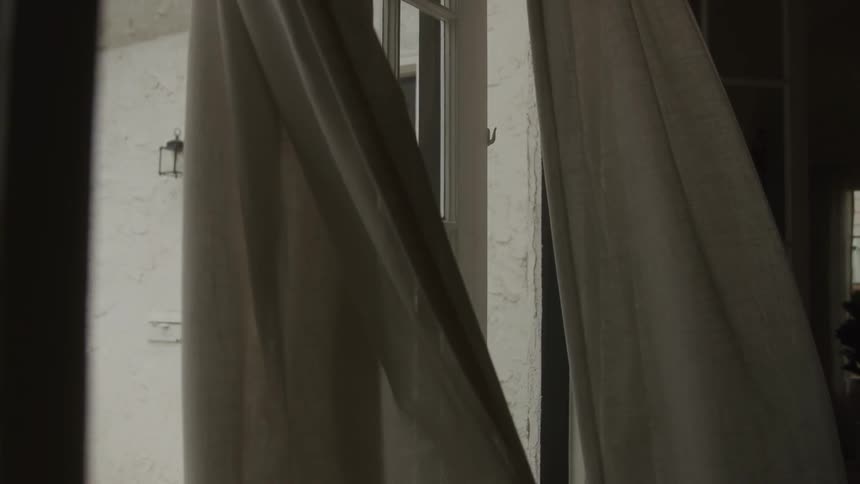 Curtains billow gently in the breeze beside a window in a softly lit interior space
