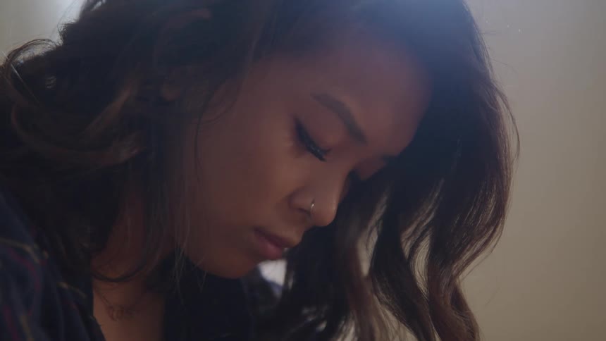 A woman with a nose ring and long hair looks down sadly while indoors