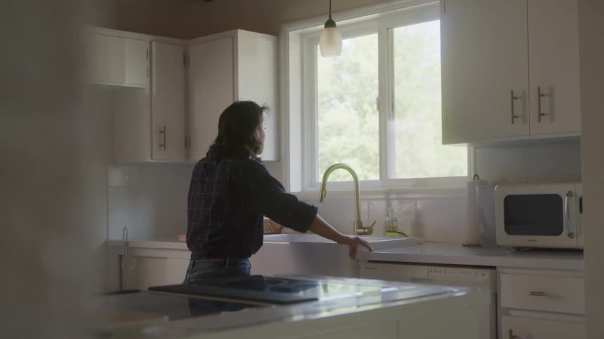 A lady stands close to the kitchen faucet in the morning looking out through the window