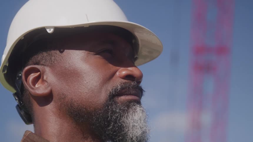 A closeup shows the face of a man in a white hard hat looking upwards with a thoughtful expression against a blue sky and a faint red structure
