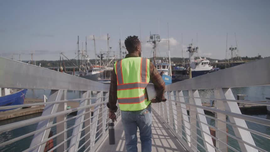 A man in a reflective safety vest walks down a dock ramp toward a harbor filled with fishing boats