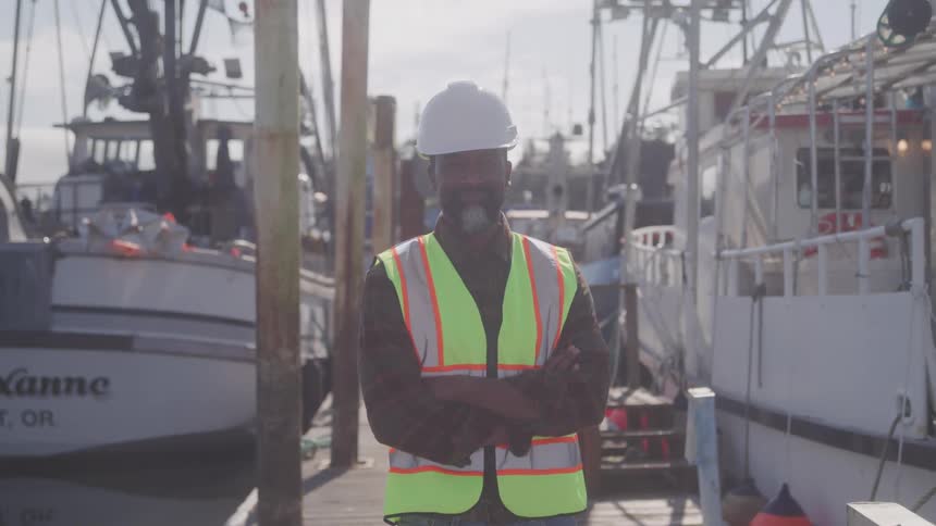 Man wearing a hardhat and a safety vest stands near a boat on a dock on a sunny day