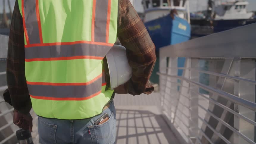 A construction worker in a safety vest walks down a dock toward boats carrying a hard hat in one hand