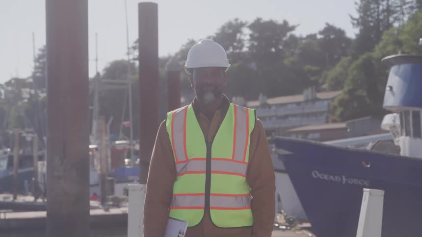 Male worker wearing a hardhat holds a clipboard while standing on the dock near a boat on a sunny day