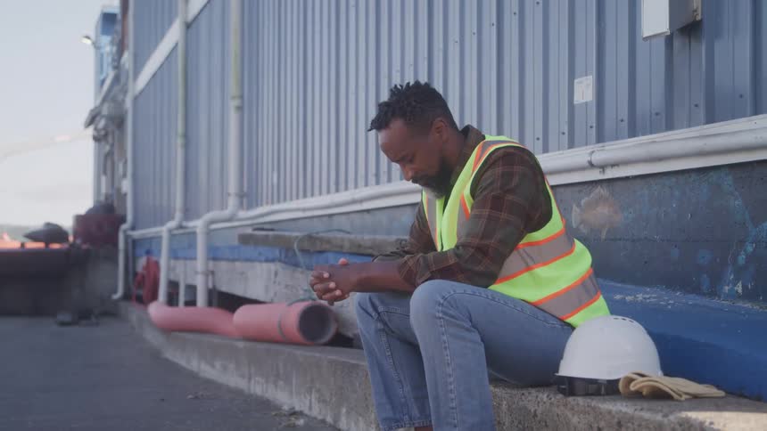 A man sits on a concrete ledge beside a warehouse at midday deep in thought wearing a green vest with a hard hat by his side