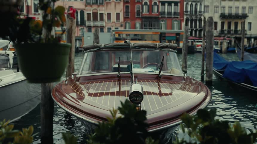A sleek wooden boat is docked on a venetian canal framed by plants and historic buildings