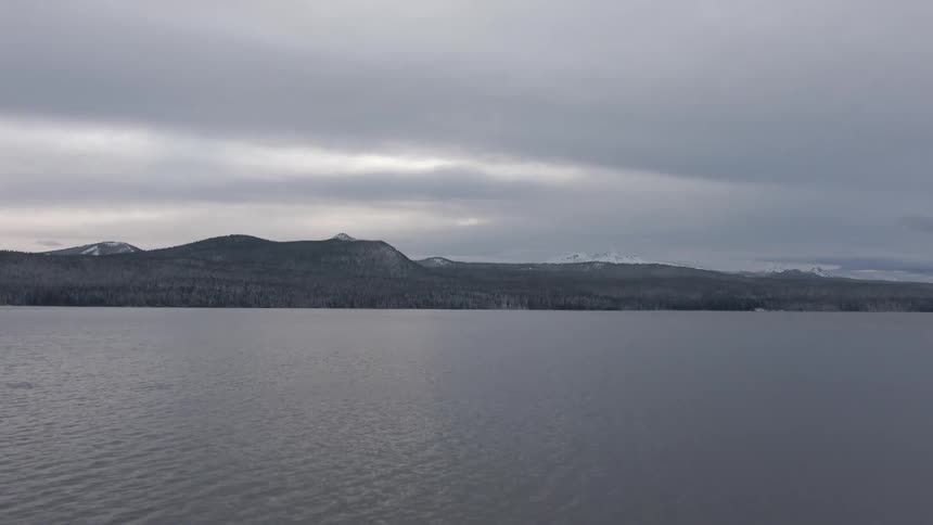 A wide shot captures a calm overcast lake with a forested mountain range in the distance beneath a gray moody sky
