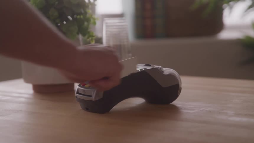 A person swipes a credit card on a card machine placed on a wooden table
