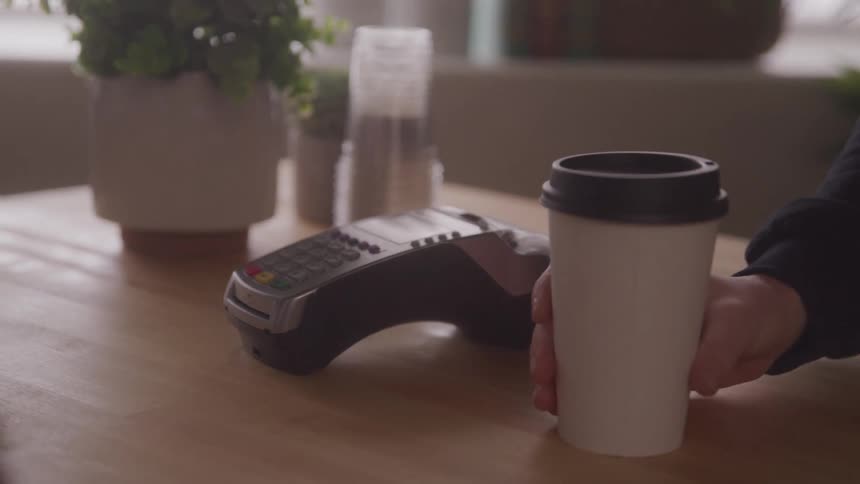 A person taps their phone on a card machine then another person places a coffee cup on the table