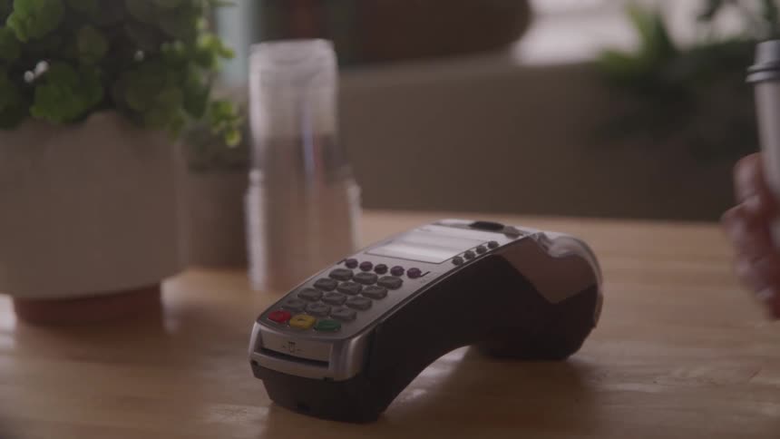 A person completes a contactless payment using a smartphone at a counter and picks up a coffee cup
