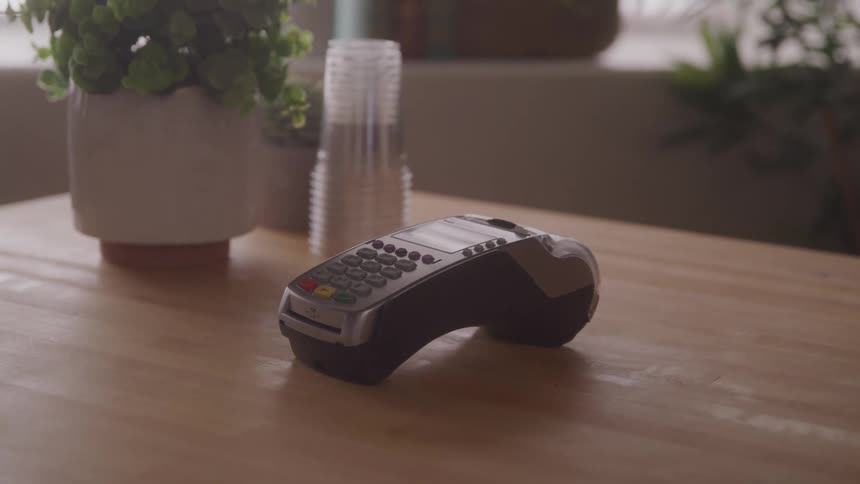 A credit card terminal on a wooden counter with cups and a plant nearby