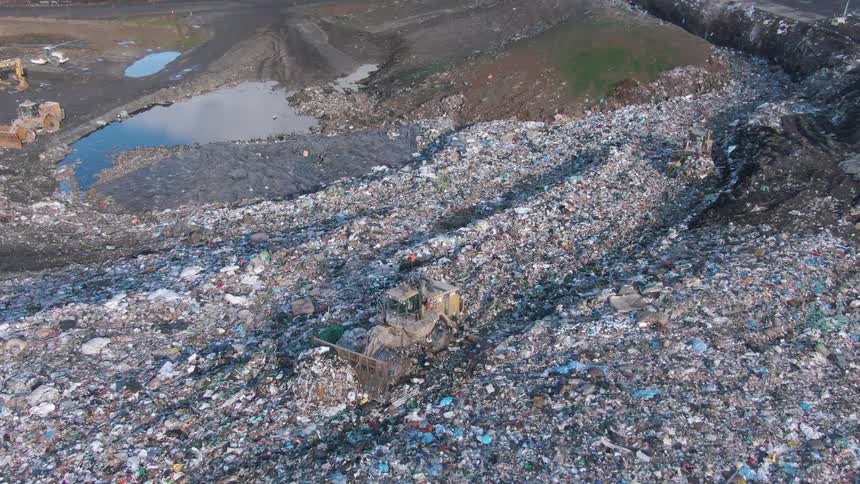 Aerial view of a landfill with bulldozers moving through large piles of garbage near a water pit