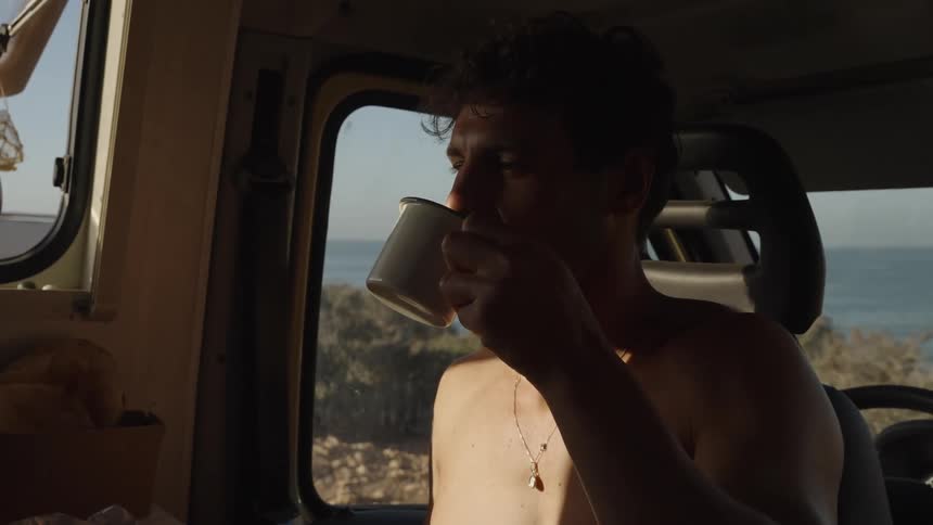 A shirtless man sips a drink in a sunlit van with an ocean view