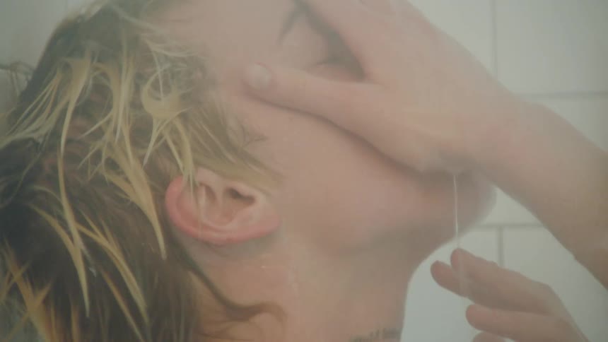 Blonde woman rinses her face and hair in a white tiled shower