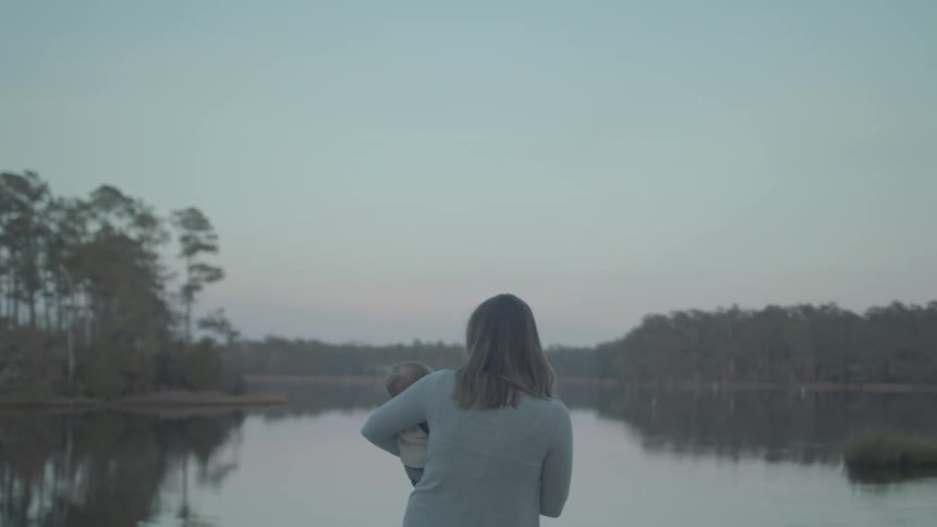Mother with a baby in her arms in front of a lake with a forest on the shore