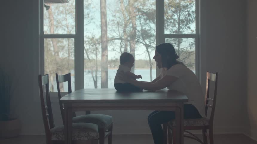 At midday a mother sits on a chair by a window with trees visible outside holding her infant son perched on the dining table and playfully teasing him