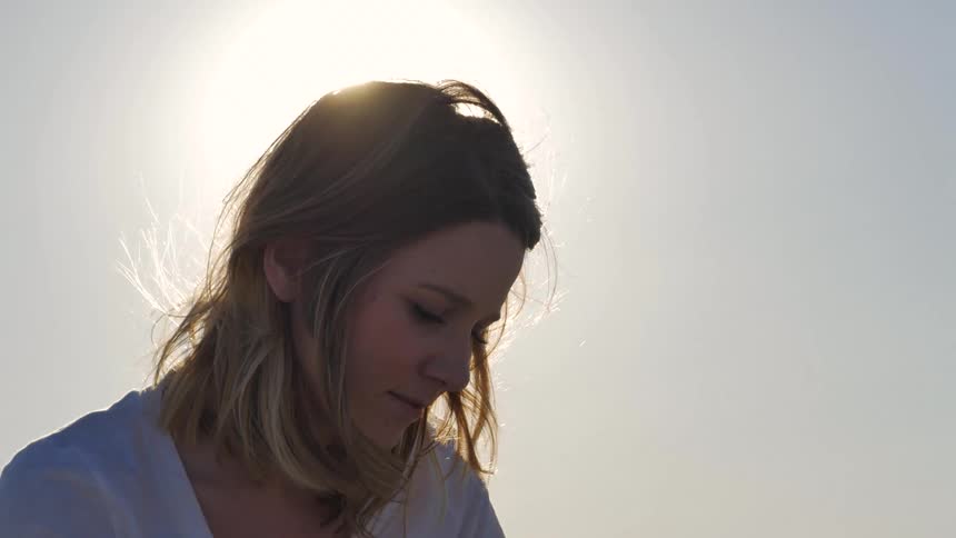 A womans face is shown in closeup backlit by the bright sun as she looks down with a gentle smile