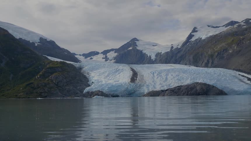 A vast glacier meets the calm waters of a lake or fjord nestled between snowcapped mountains under a cloudy sky