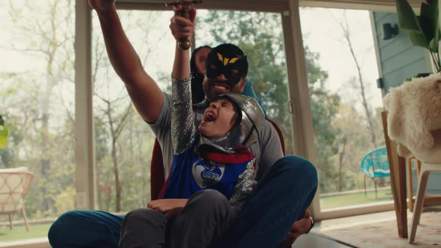 A family in costumes is laughing and playing pretend in a bright room with large windows