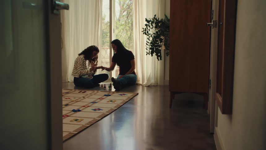 A teenage girl sits on the floor in a room with large windows and a plant carefully painting a womans nails beside a patterned rug