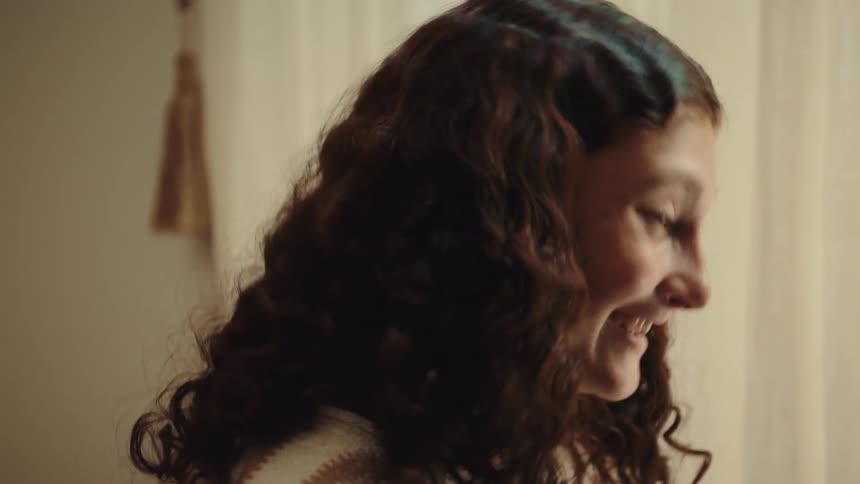 A teenage girl laughs and chats with someone offcamera bathed in warm indoor light