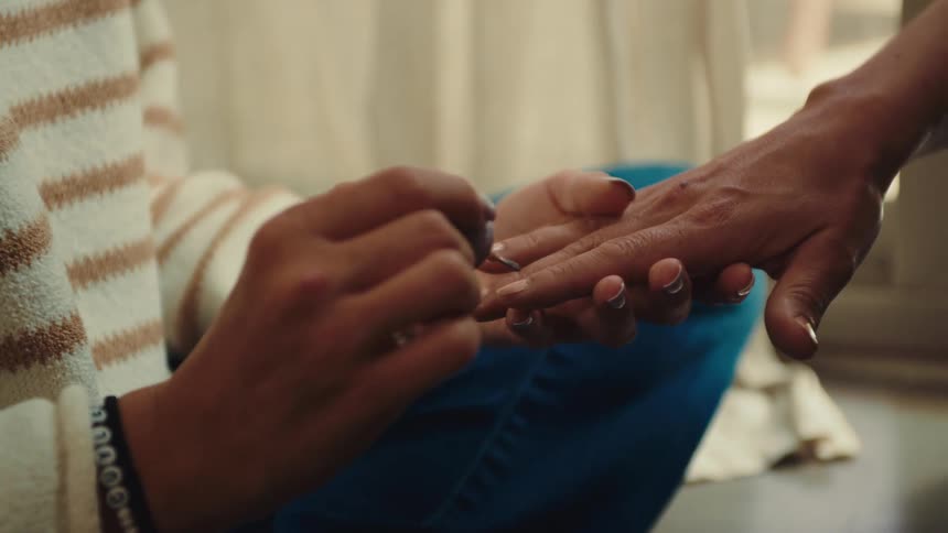 A woman applies nail polish to another womans fingers while they sit on the floor at midday