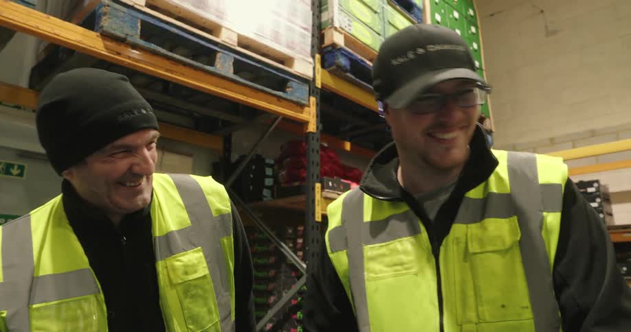 Two male workers wearing green vests and hats are standing in a warehouse engaged in a lively conversation and laughing together
